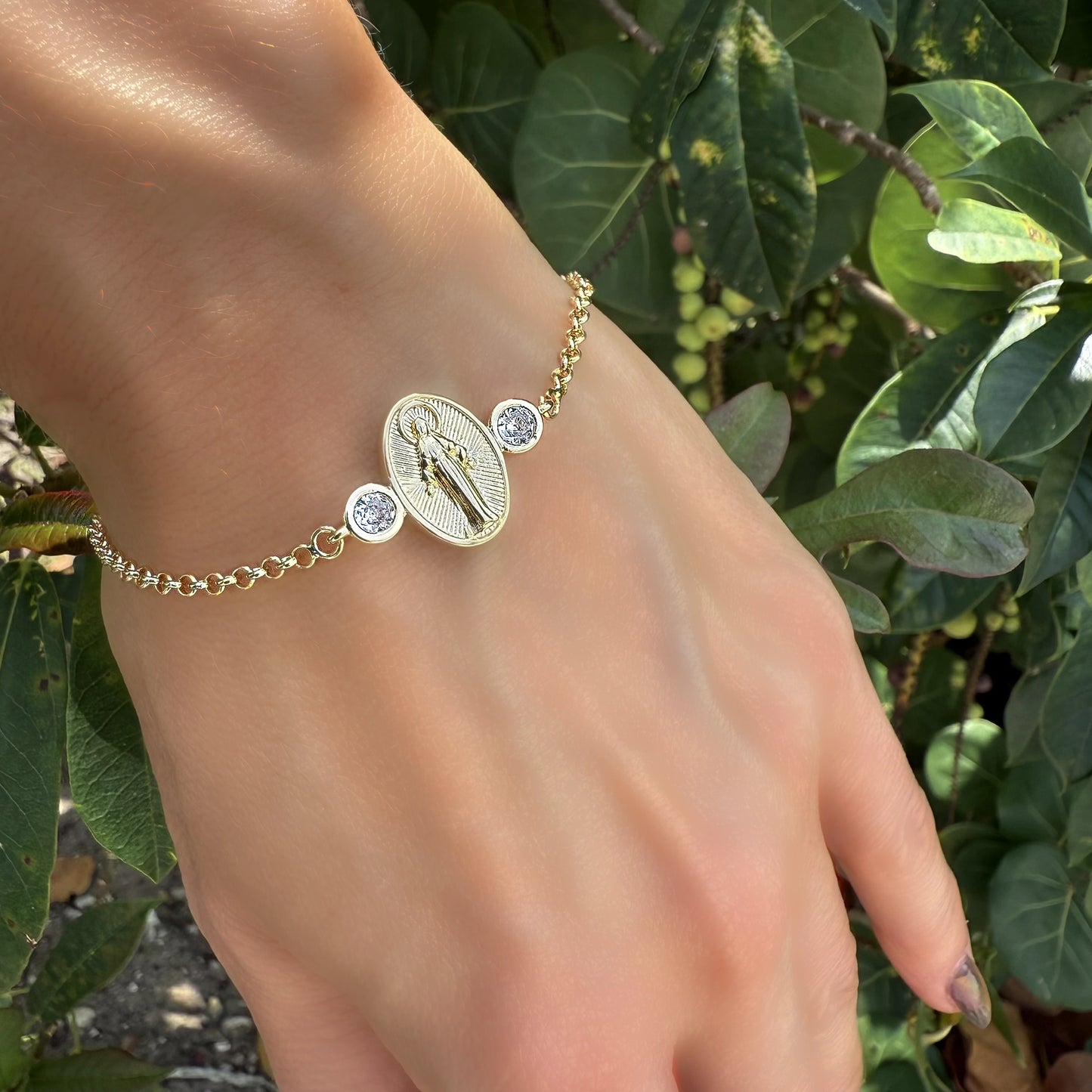 18k gold filled Virgin Mary medallion bracelet featuring two bezel-set CZ accents, worn on a hand outdoors with green foliage in the background. Symbolic and elegant Catholic jewelry perfect for daily wear.