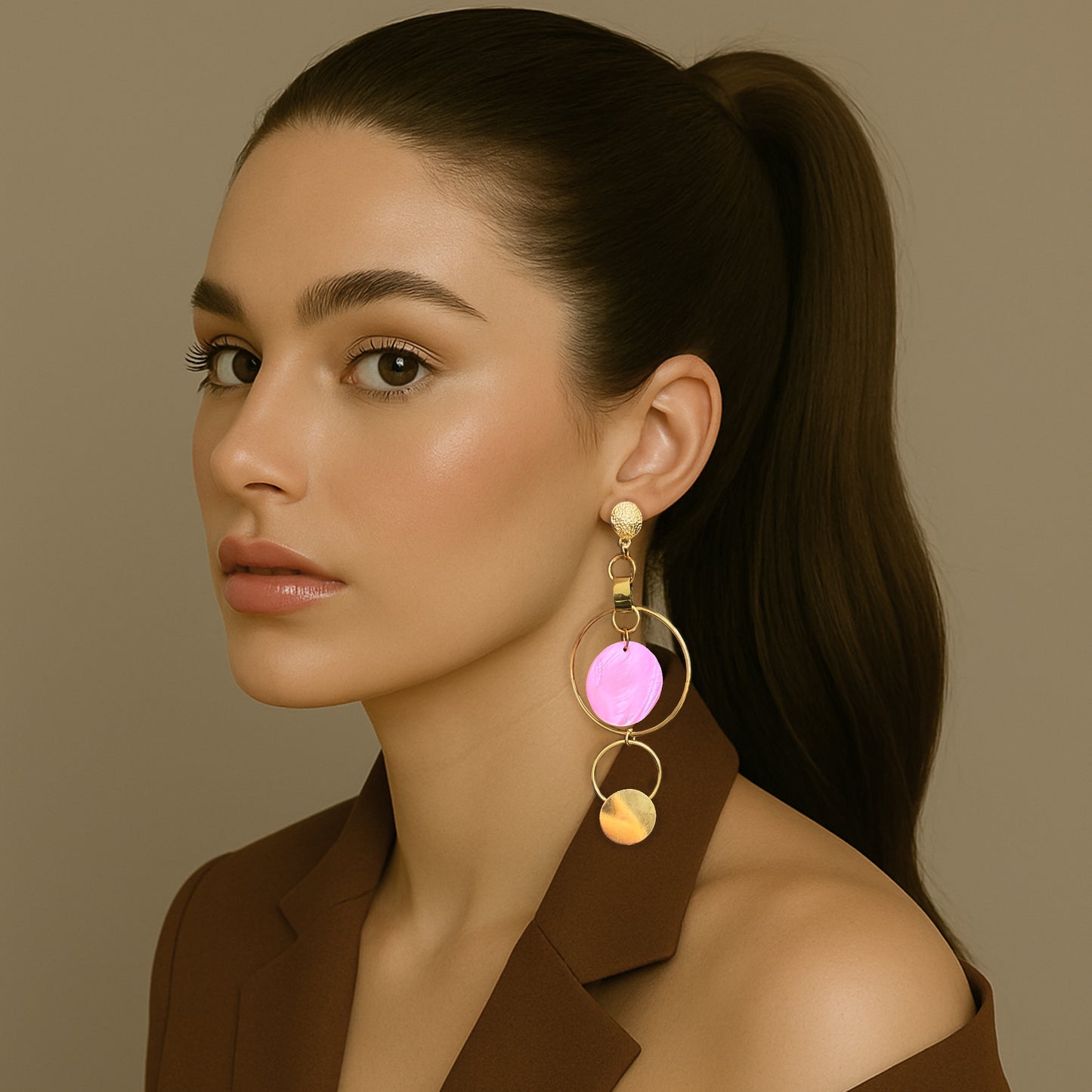 Elegant woman in a brown blazer with a sleek ponytail wearing bold asymmetric gold hoop earrings featuring a pink shell disc and hammered metal accents.