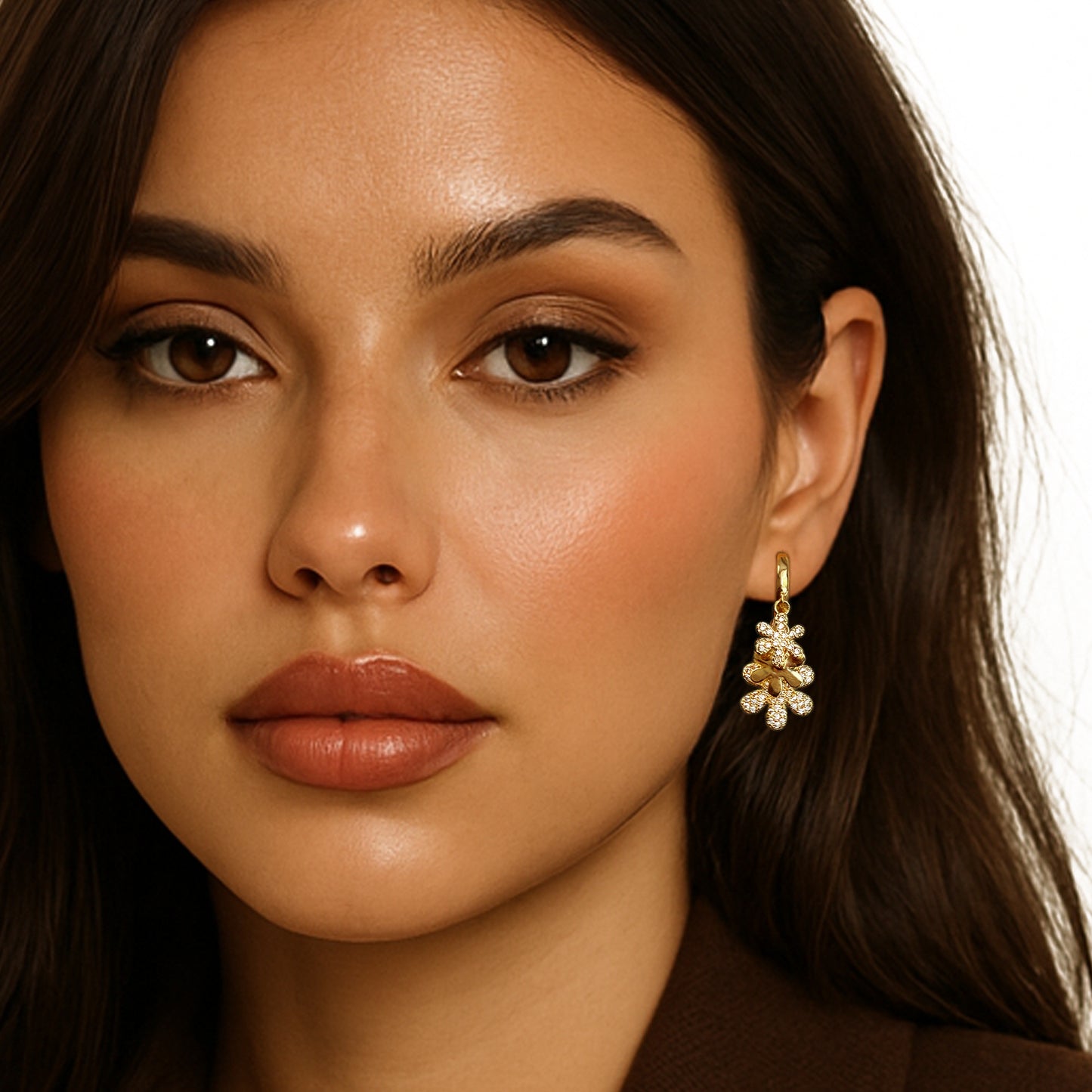 Close-up of a woman wearing gold huggie hoop earrings adorned with dangling cubic zirconia floral charms, styled with a brown blazer and warm-toned makeup for a polished, elegant appearance.