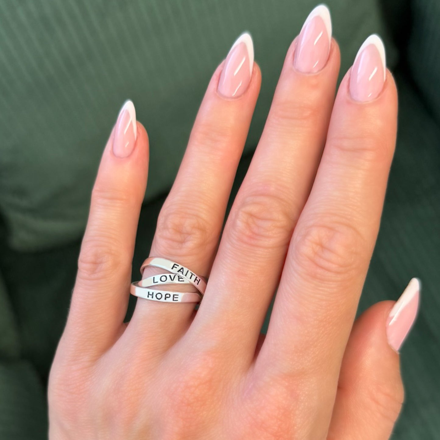 Three-band inspirational ring set in sterling silver reading "FAITH", "LOVE", and "HOPE" on a woman's hand with French tip nails.