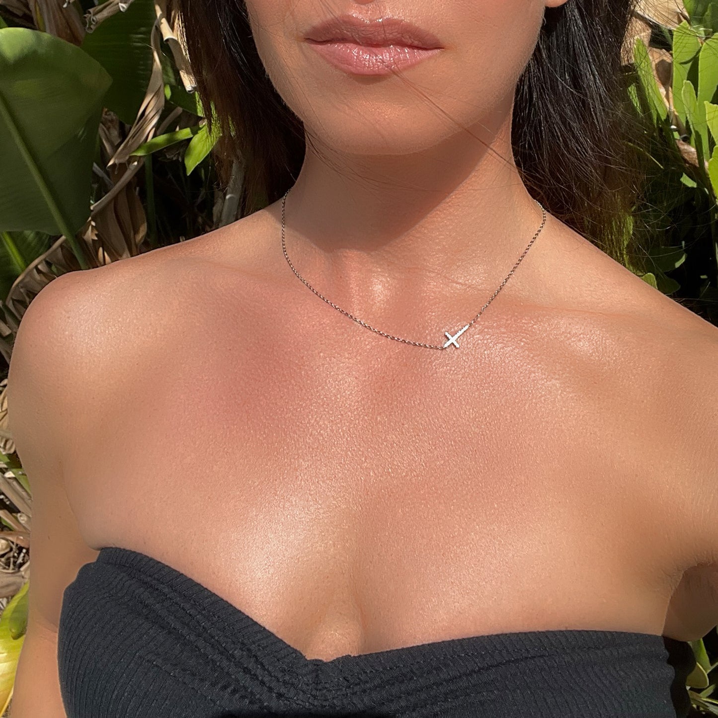 Sterling silver sideways cross necklace with cubic zirconia, modeled outdoors on a woman in a strapless black top, emphasizing neckline and dainty chain.