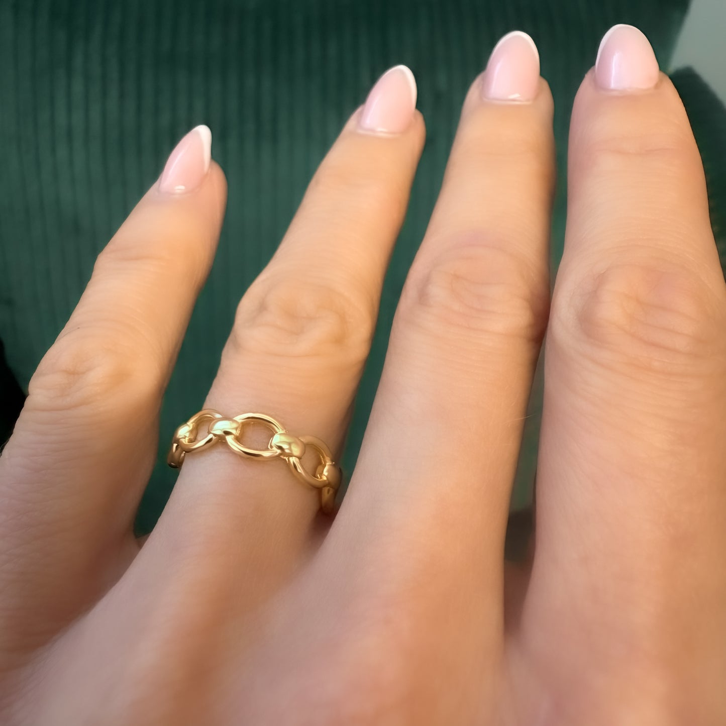 Close-up of a woman's hand wearing a gold chain ring with French manicure, showcasing the delicate link design against a soft green background.