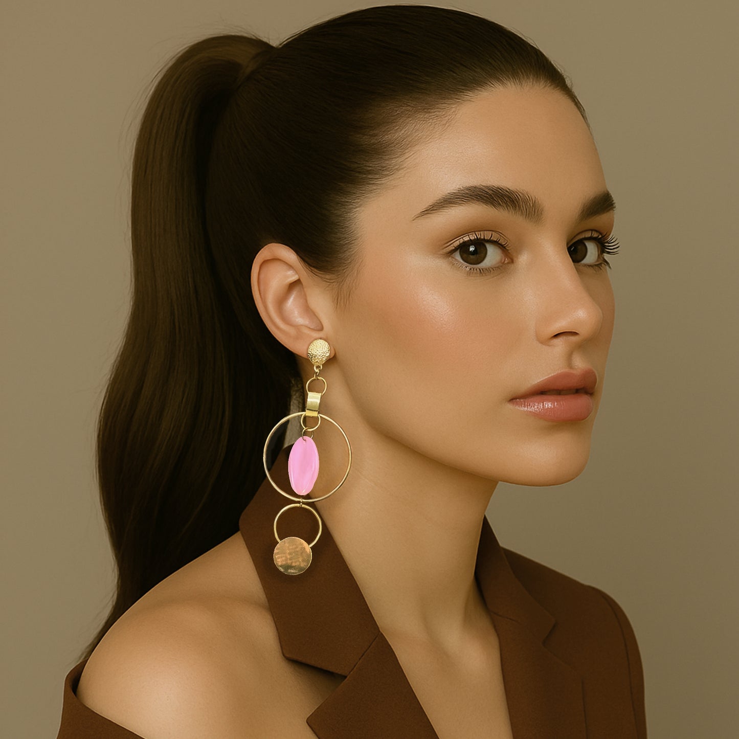 Elegant woman in a brown blazer with a sleek ponytail wearing bold asymmetric gold hoop earrings featuring a pink shell disc and hammered metal accents.