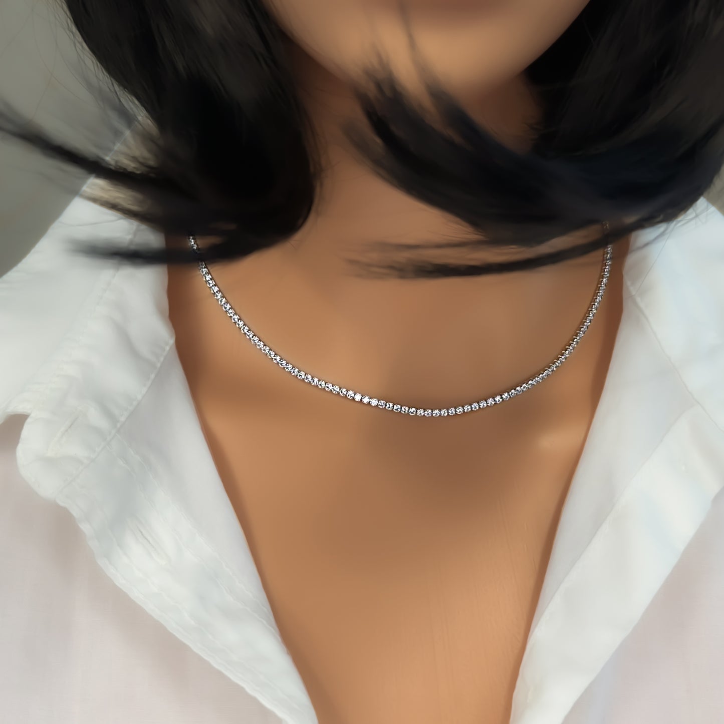 Close-up of a model wearing a classic sterling silver tennis necklace with a continuous line of round-cut cubic zirconia stones. The necklace rests elegantly along the collarbone and is styled with an open white shirt and soft, flowing hair.