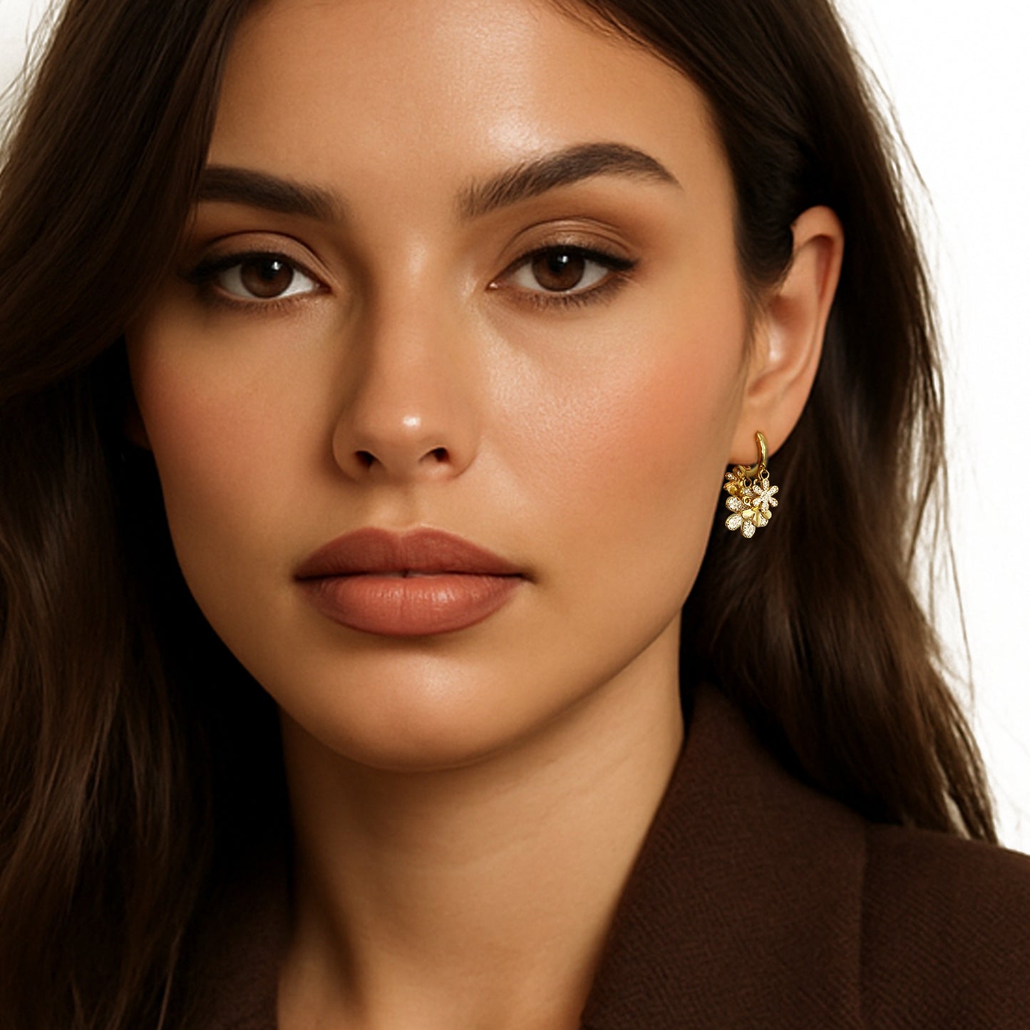 Close-up of a woman wearing gold huggie hoop earrings with dangling cubic zirconia flower charms, styled with a brown blazer and soft neutral makeup for an elegant, feminine look.