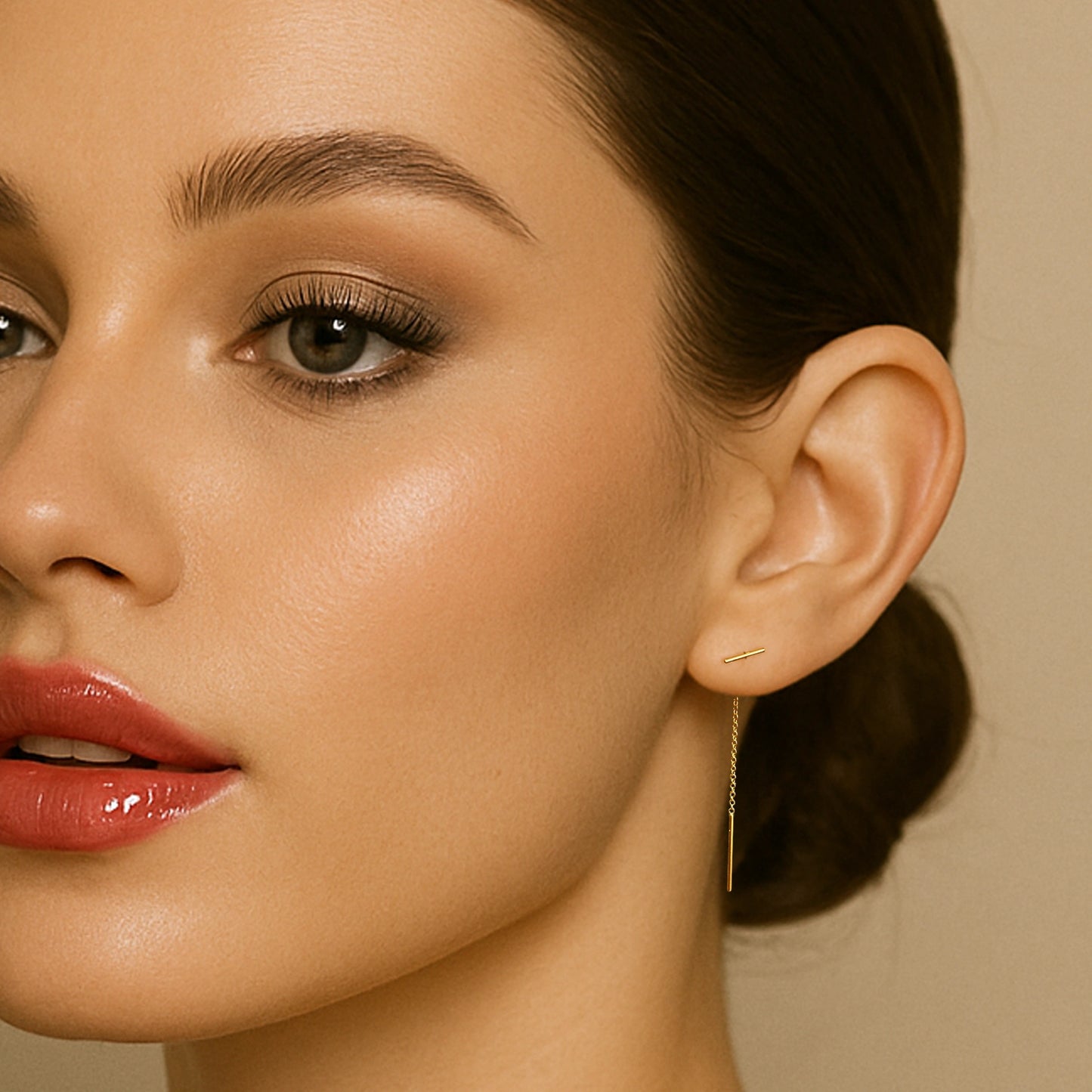 Close-up of a woman wearing delicate gold threader earrings with a sleek t-bar drop, styled with soft glam makeup and a low bun hairstyle.