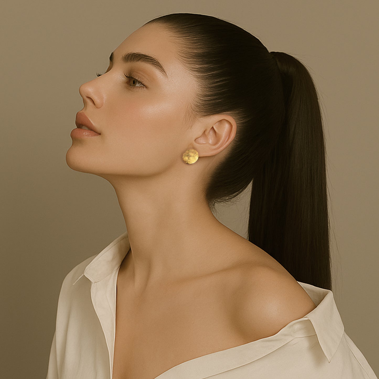Elegant woman in an off-shoulder white blouse wearing simple round gold disc stud earrings, with her head tilted upward and hair in a sleek ponytail.