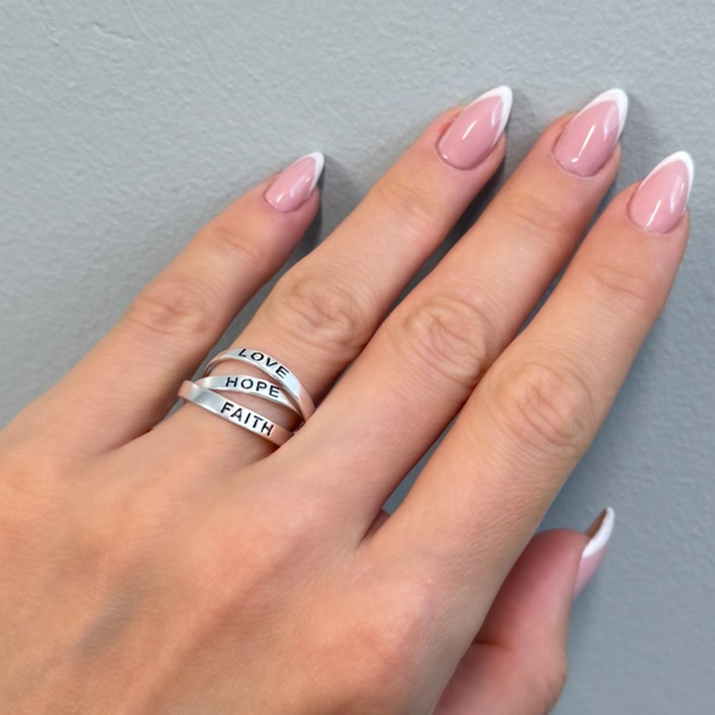 Sterling silver ring stack engraved with "LOVE", "HOPE", and "FAITH" worn on manicured hand against gray background.
