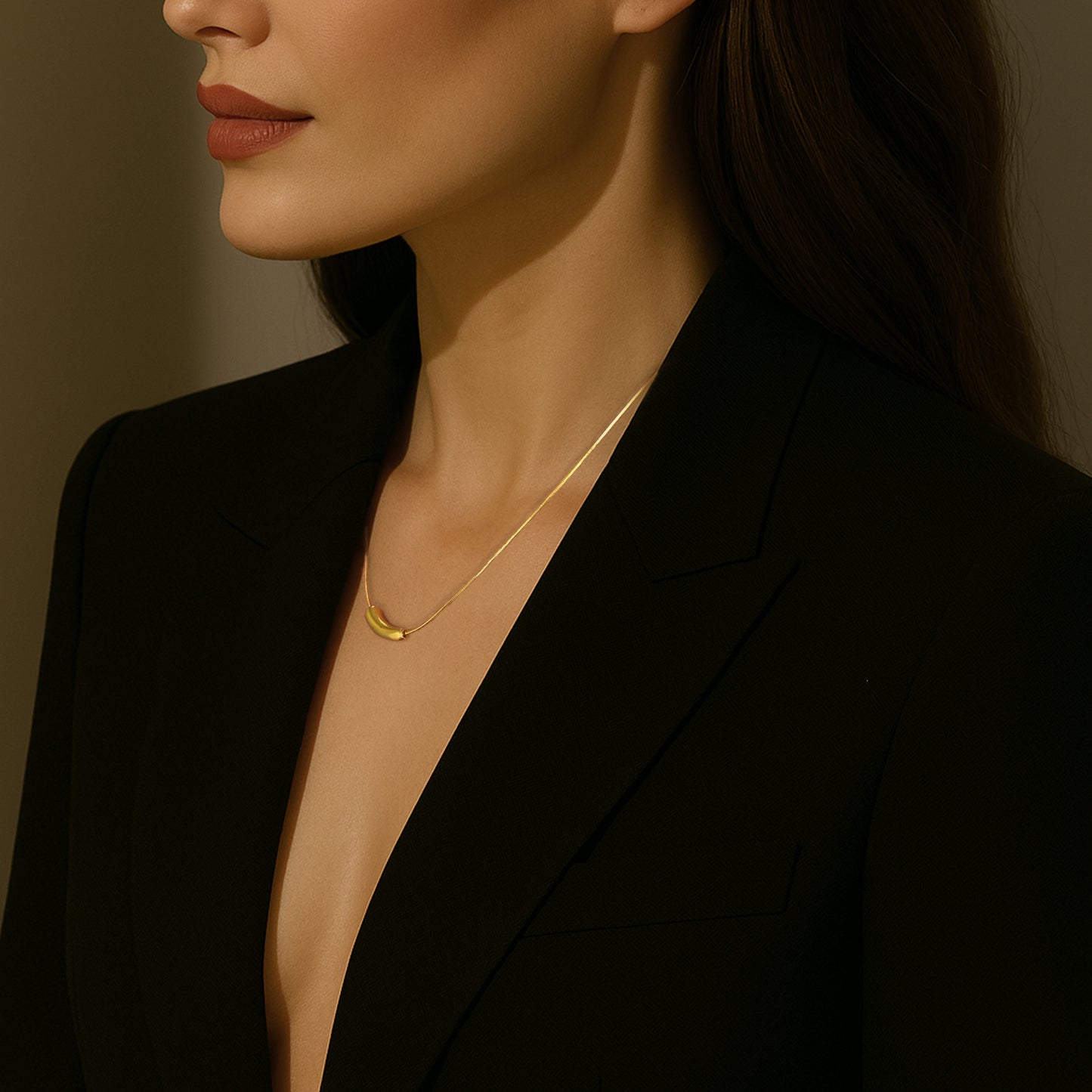 A close-up of a woman wearing a black blazer and a dainty gold necklace with a curved bar pendant on a snake chain. Her face is partially visible in profile, with warm lighting casting soft shadows against a neutral background.