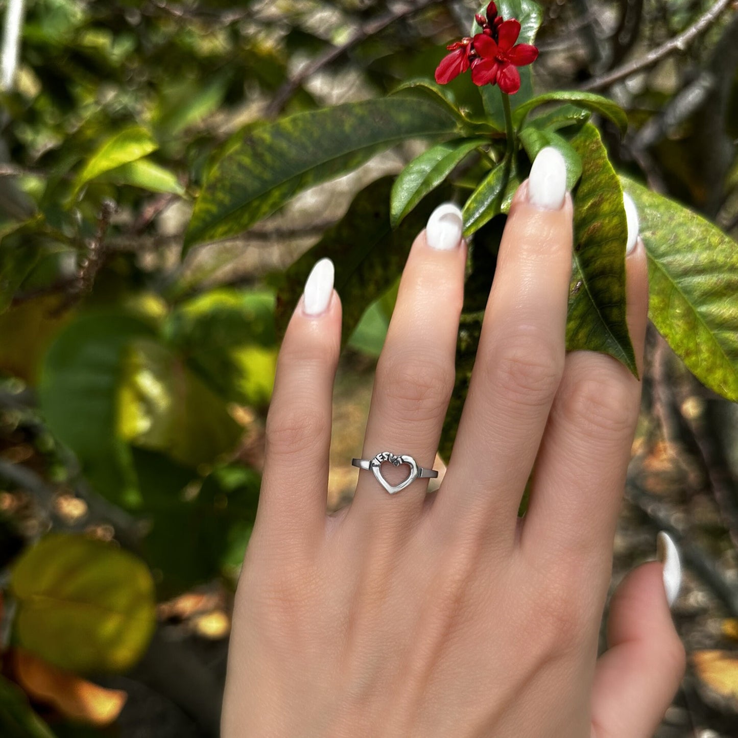 A sterling silver ring featuring an open heart engraved with “Jesus,” worn on a hand with white nails against a leafy green background.