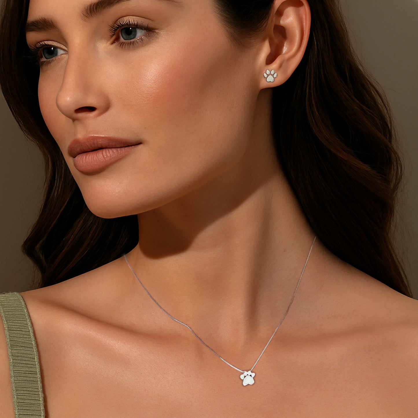 Model wearing a dainty sterling silver paw print necklace and matching paw print stud earrings, styled with natural makeup and a green ribbed tank top.