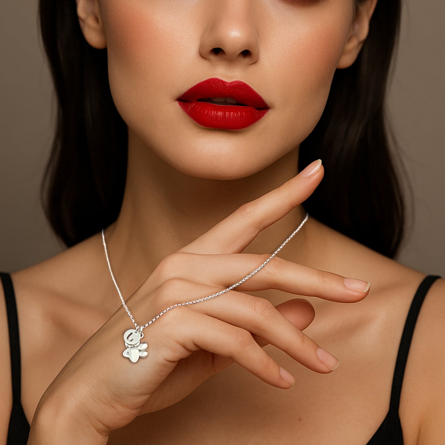 Close-up of a woman with bold red lips holding a delicate sterling silver necklace featuring a paw print charm and a personalized initial pendant.