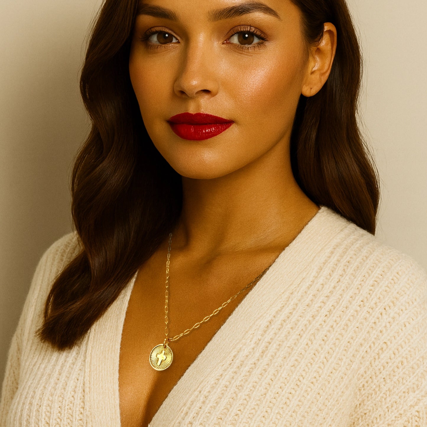 Beautiful woman with radiant skin and bold red lipstick wearing an elegant 18k gold filled cross medallion necklace over a cream knit V-neck sweater. The round pendant features a raised cross design with radiant textured detailing, offering a stylish and faith-inspired look perfect for layering.