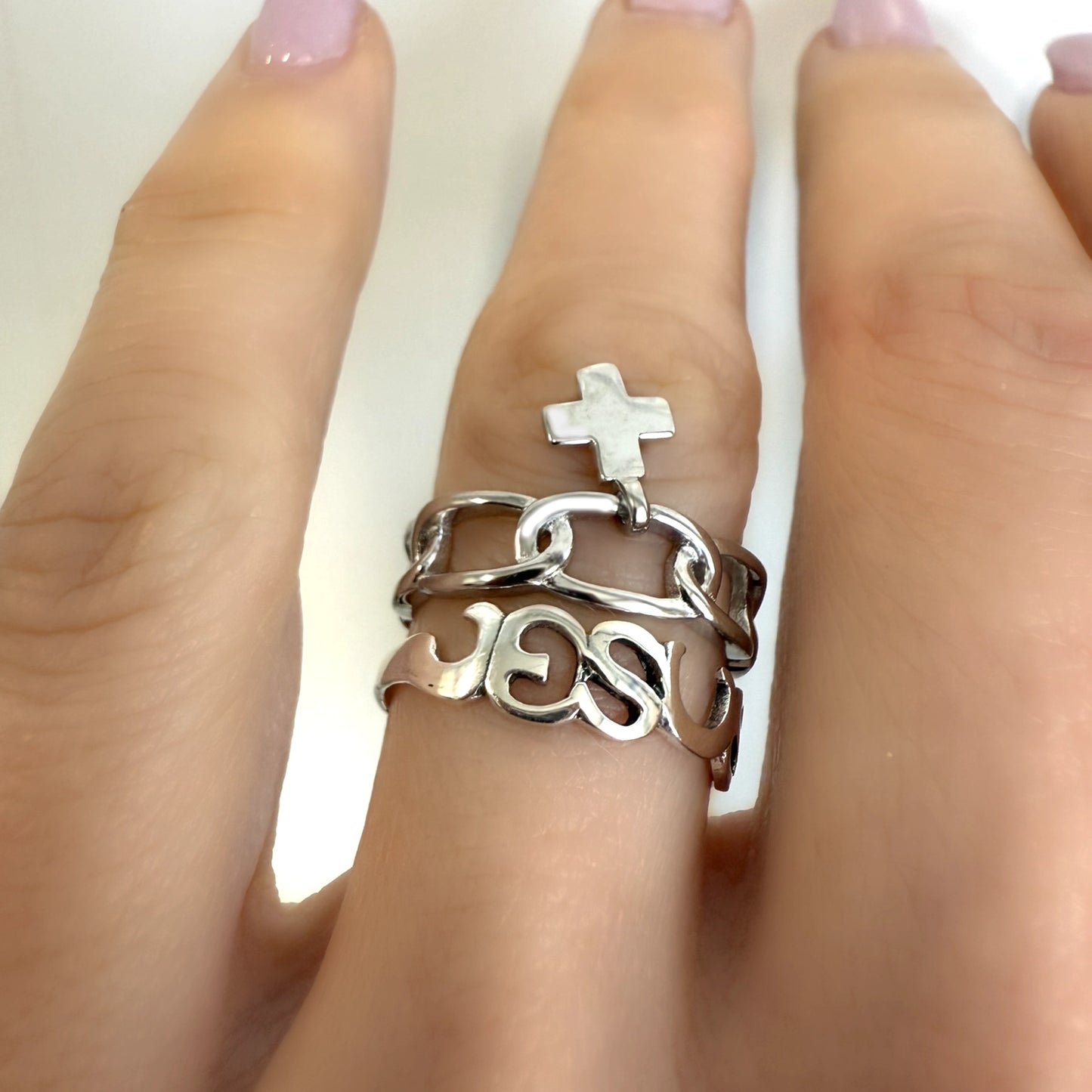 Close-up view of a sterling silver Christian ring set on finger, featuring a βJESUSβ cutout band, a chunky chain ring, and a dangling cross charm.