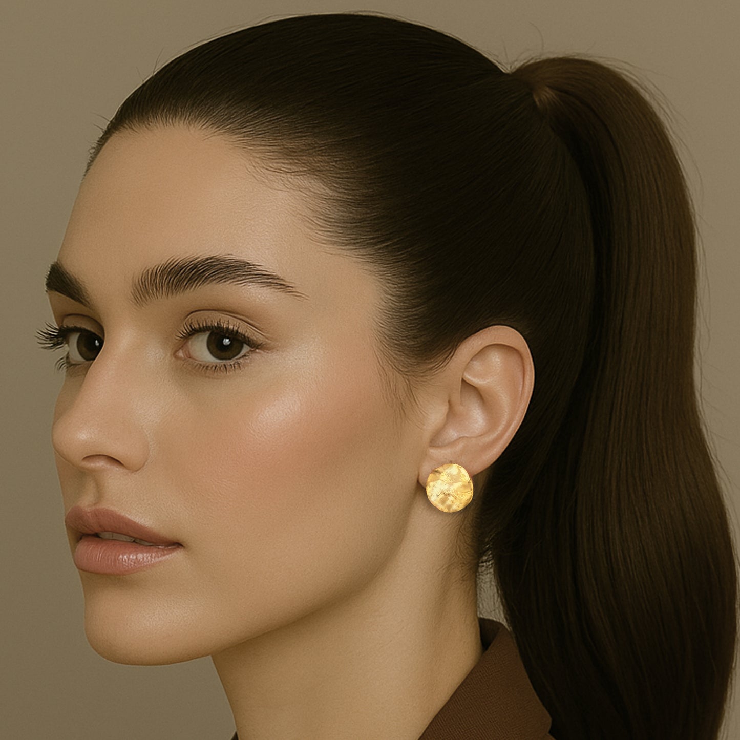 Close-up of a woman with a sleek ponytail wearing minimalist round gold disc stud earrings, styled with a brown blazer and natural makeup.
