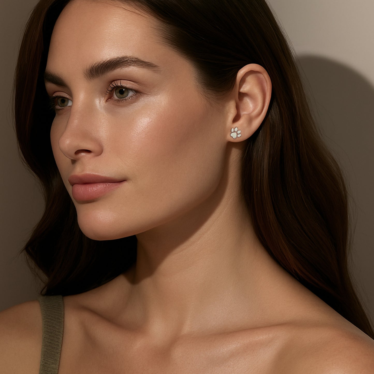 A woman with long brown hair and soft makeup is shown in side profile wearing a small silver paw-shaped stud earring. She is dressed in a green tank top, and the lighting creates gentle shadows against a neutral background, emphasizing her natural beauty and the earring.