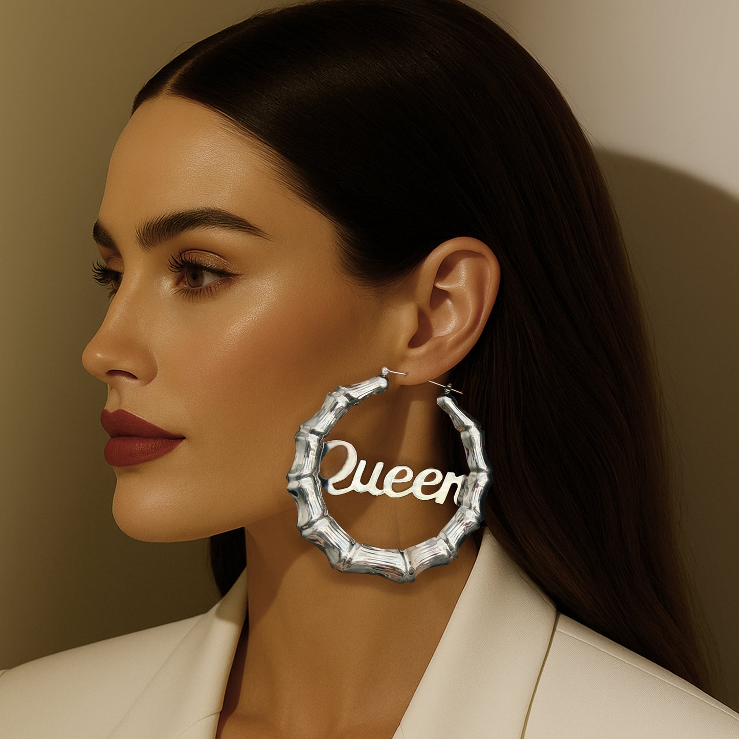 Side profile of a woman wearing oversized silver-tone bamboo hoop earrings with the word "Queen" in bold script, styled with sleek hair and bold makeup.