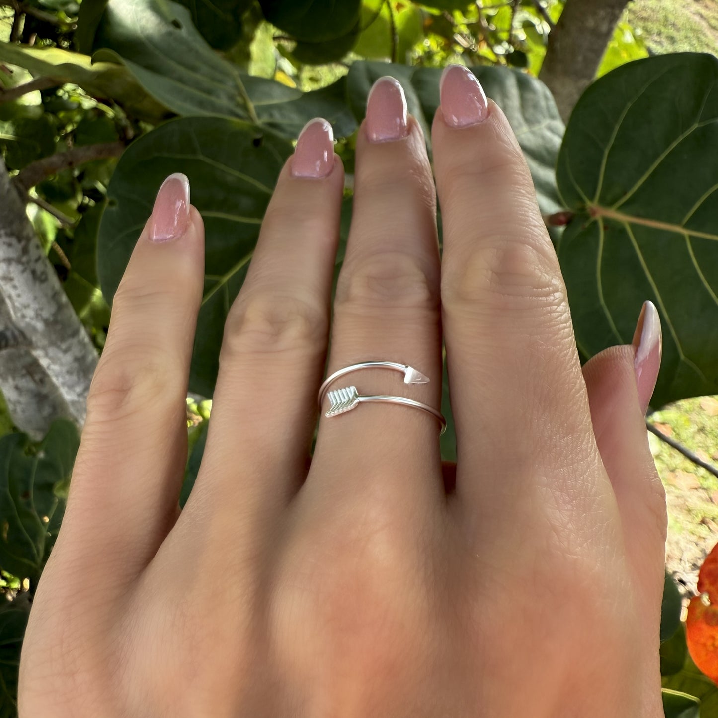Full-hand view of a delicate silver arrow ring with a modern wrap-around shape, worn on the middle finger with glossy nude-pink manicure in a leafy outdoor setting.
