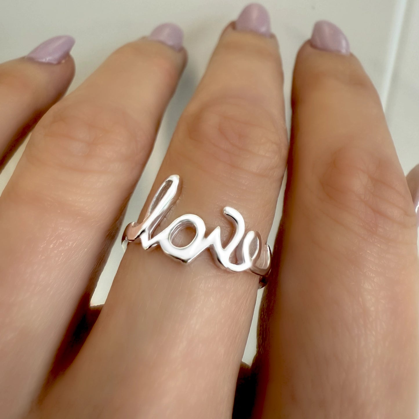 Sterling silver cut-out ring spelling the word "love" in a cursive script, worn on a hand with light pink polished nails.