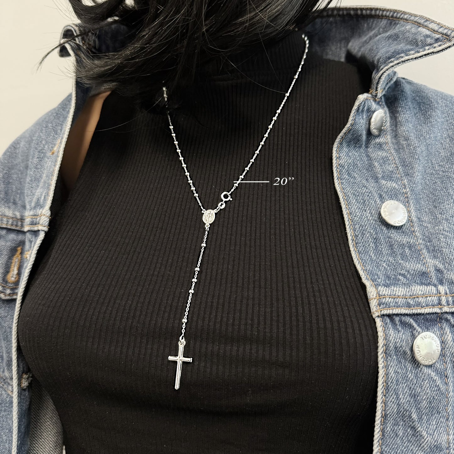 Model wearing a 20-inch sterling silver rosary bead necklace with a Virgin Mary connector and vertical cross pendant. Styled over a black ribbed turtleneck and layered with a denim jacket for a modern, faith-inspired look.