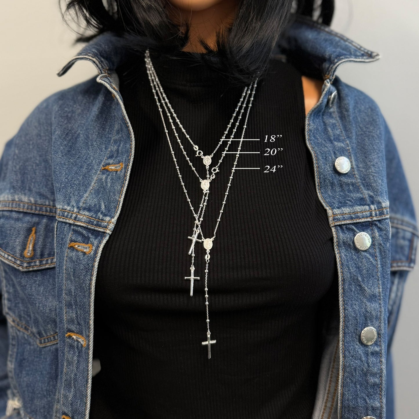 Model layered in 18", 20", and 24" sterling silver rosary bead necklaces, each featuring a Virgin Mary connector and drop cross pendant. Perfect for layering or choosing your ideal fit, shown with a denim jacket and black ribbed top.