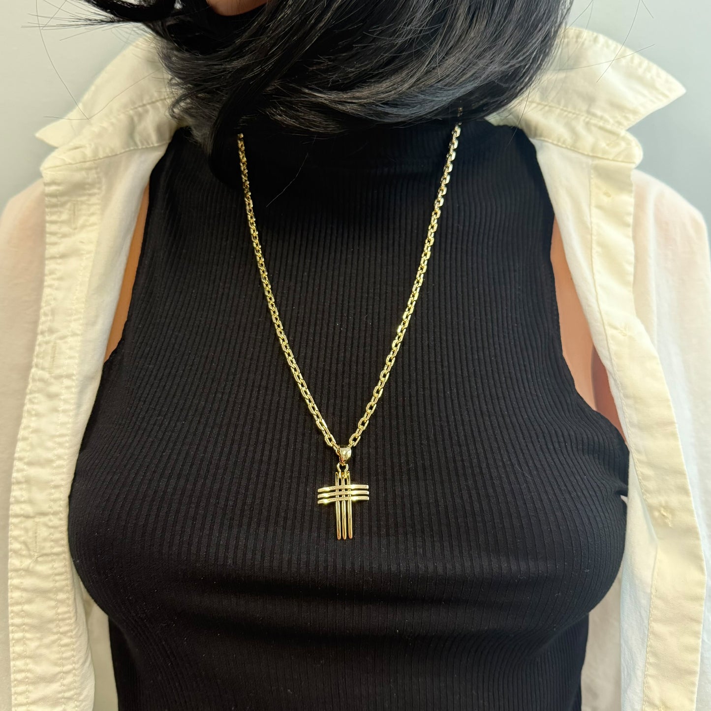 Front-facing view of a women dressed in a black ribbed top and open cream shirt, showcasing a statement 18k gold filled cross necklace with a thick chain and sculptural, layered cross pendant.