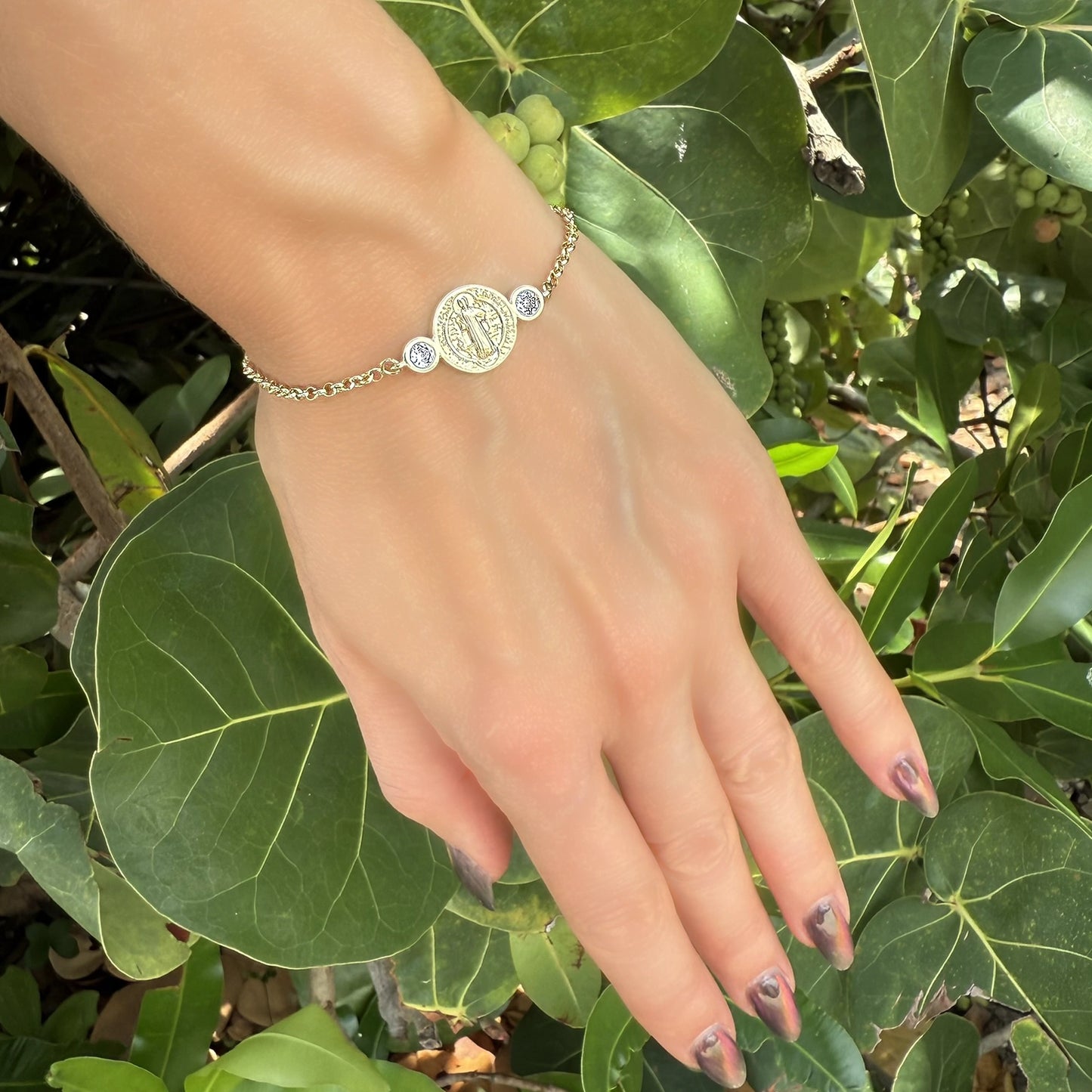 18K gold-filled Saint Benedict medallion bracelet featuring bezel-set CZ stones, worn on the wrist with leafy green foliage as a backdrop.