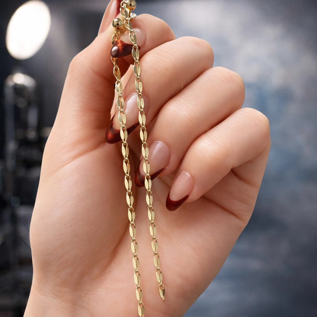 Close-up square photo of elegant gold chain drop earrings held in a woman’s hand, smooth youthful skin, refined burgundy manicure, sharp focus and contrast, professional studio lighting, soft blurred modern background, luxury jewelry product photography.