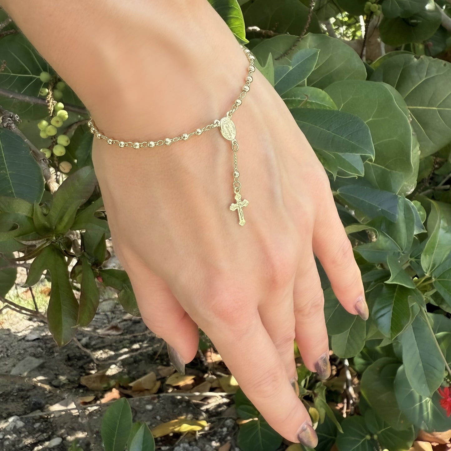 Hand wearing an 18k gold filled beaded rosary bracelet outdoors, featuring a dangling sculpted cross and Virgin Mary medallion charm, with a natural green foliage background.