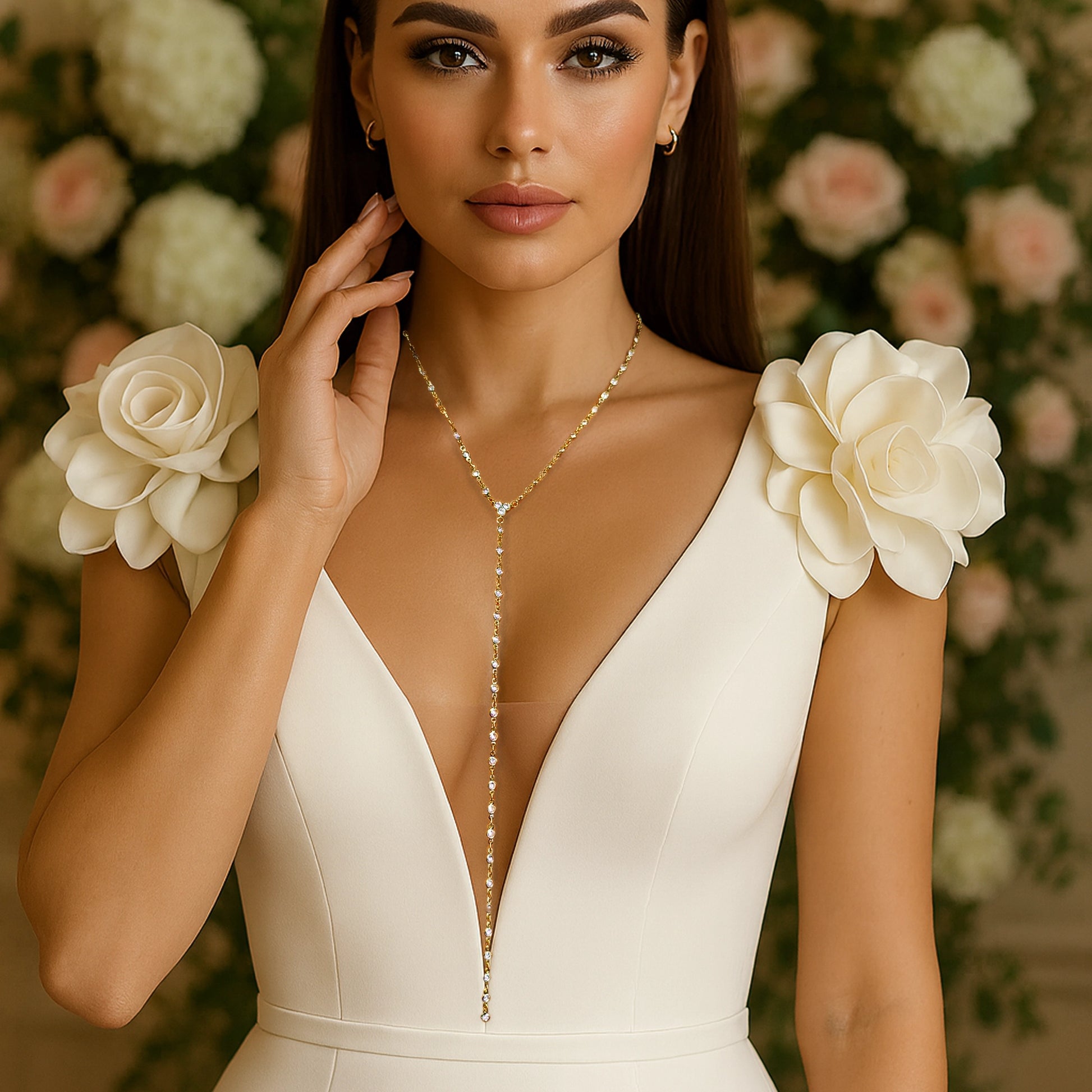 Elegant bride wearing a deep V-neck ivory bridal gown adorned with large fabric flower details on the shoulders, showcasing a delicate 18K gold filled lariat necklace with sparkling CZ stones, set against a soft floral background.