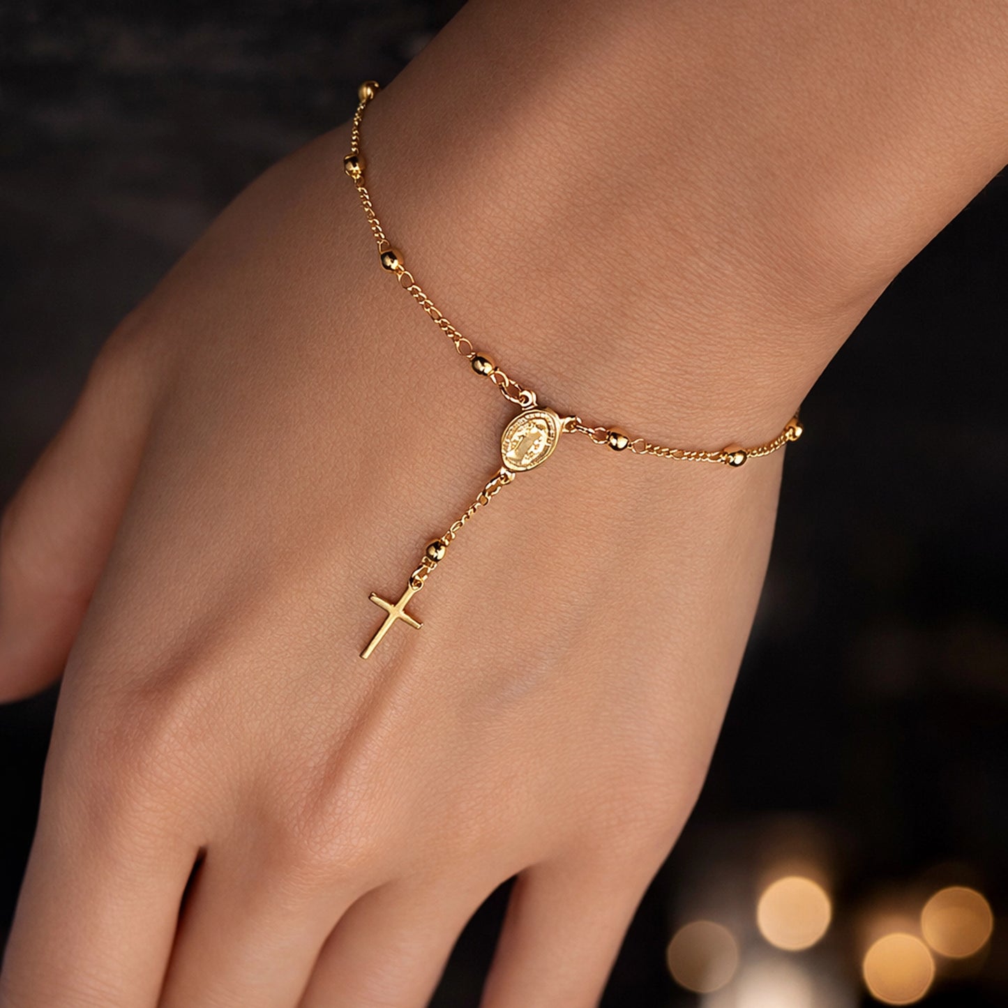 Gold religious hand bracelet with cross charm and Virgin Mary medallion on delicate bead chain, close-up lifestyle studio photo with soft warm lighting, polished golden tone, and focus on elegant Catholic jewelry detail.