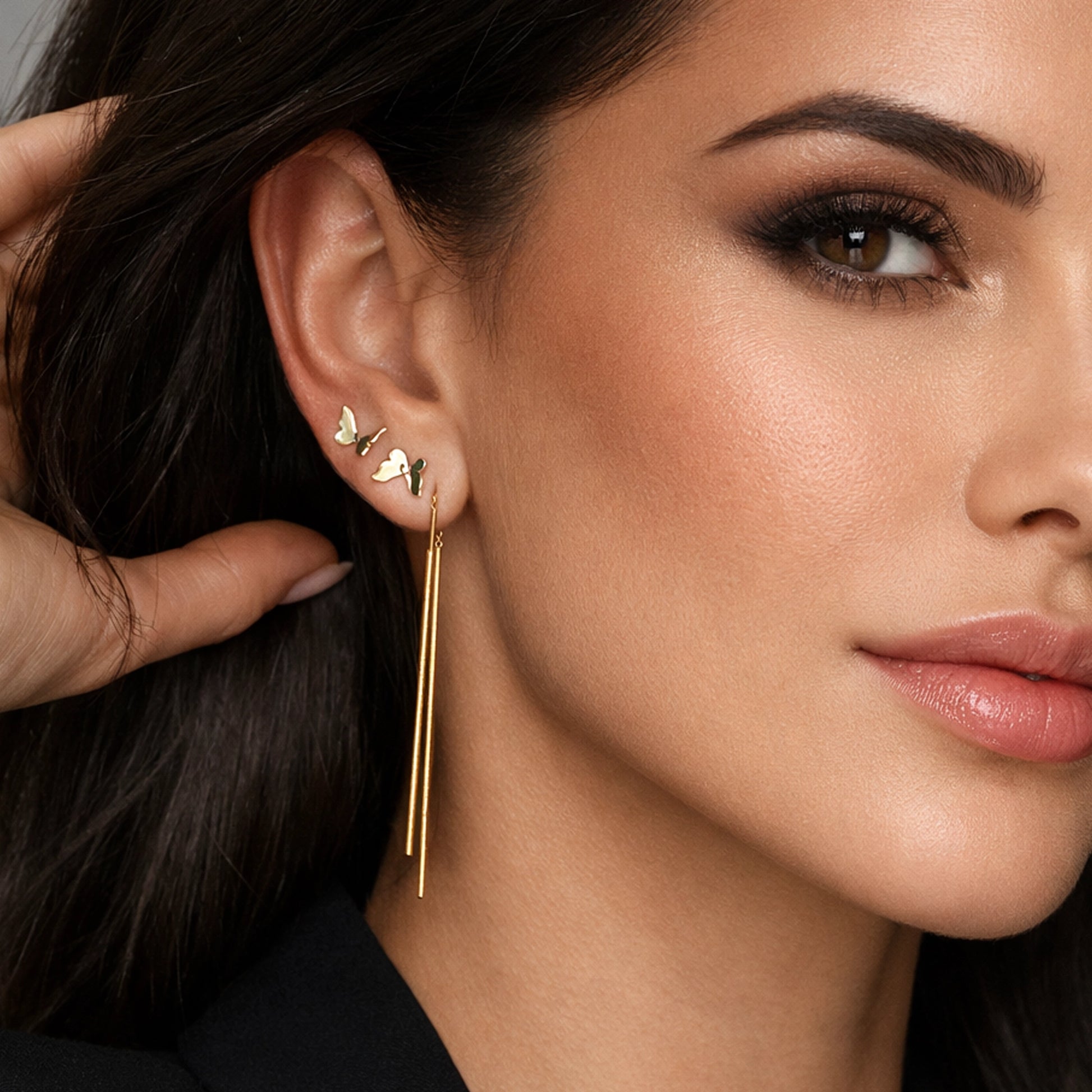 Close-up portrait of a brunette woman wearing mismatched gold earrings: a long double bar dangle earring and delicate butterfly stud earrings, styled in a stacked ear look. The model has smooth, glowing skin, soft neutral makeup, and is photographed in professional studio lighting against a clean background, highlighting the elegant gold jewelry details.
