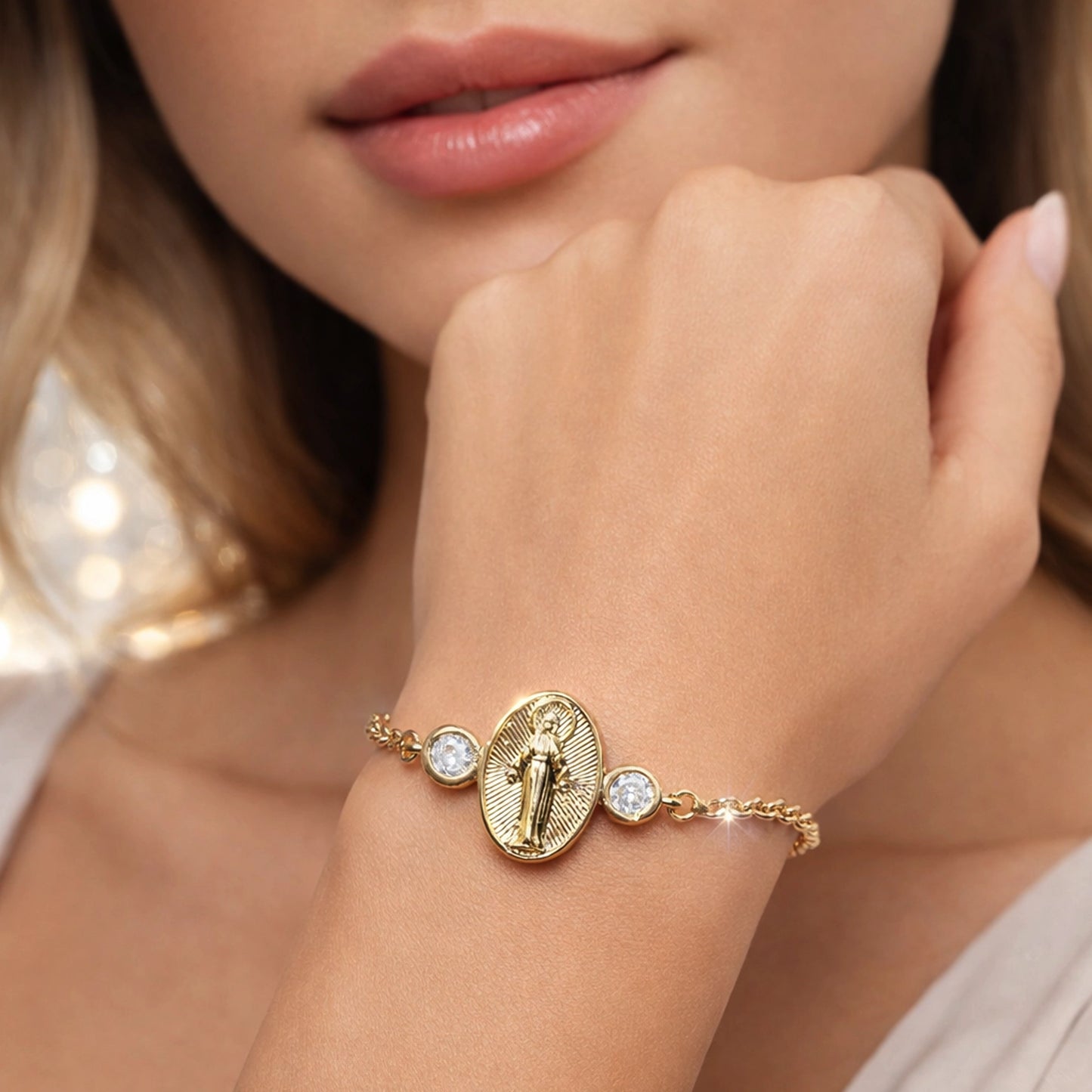 Close-up of a beautiful model holding her hand near her chin, highlighting a gold religious bracelet with an oval Virgin Mary medallion and two sparkling clear stones on a delicate chain, photographed with soft professional lighting and a light elegant background.