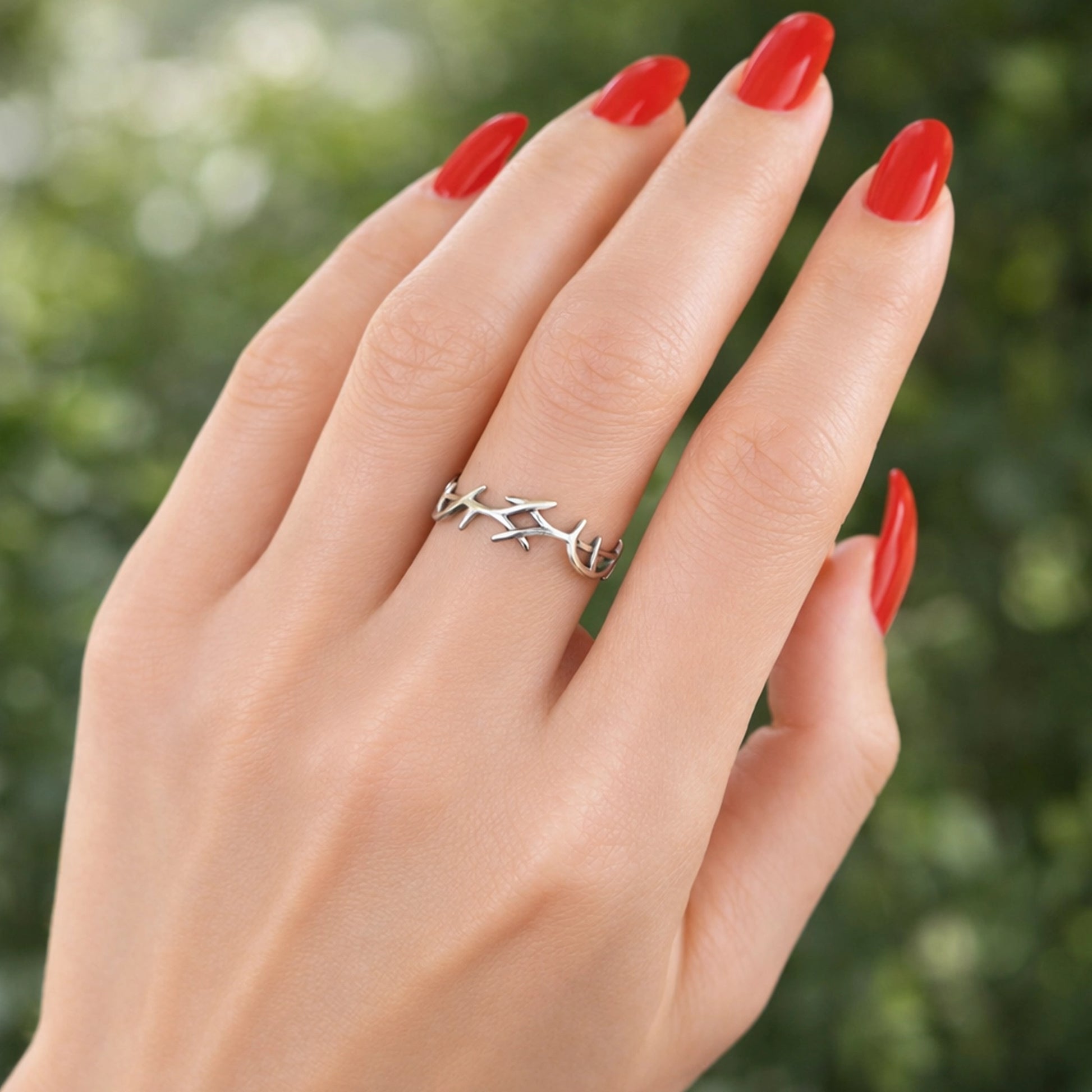Silver thorn ring wrapped around a woman’s finger, shown on a hand with glossy red manicure, soft natural skin texture, and professional blurred green background highlighting the minimalist sterling silver design.