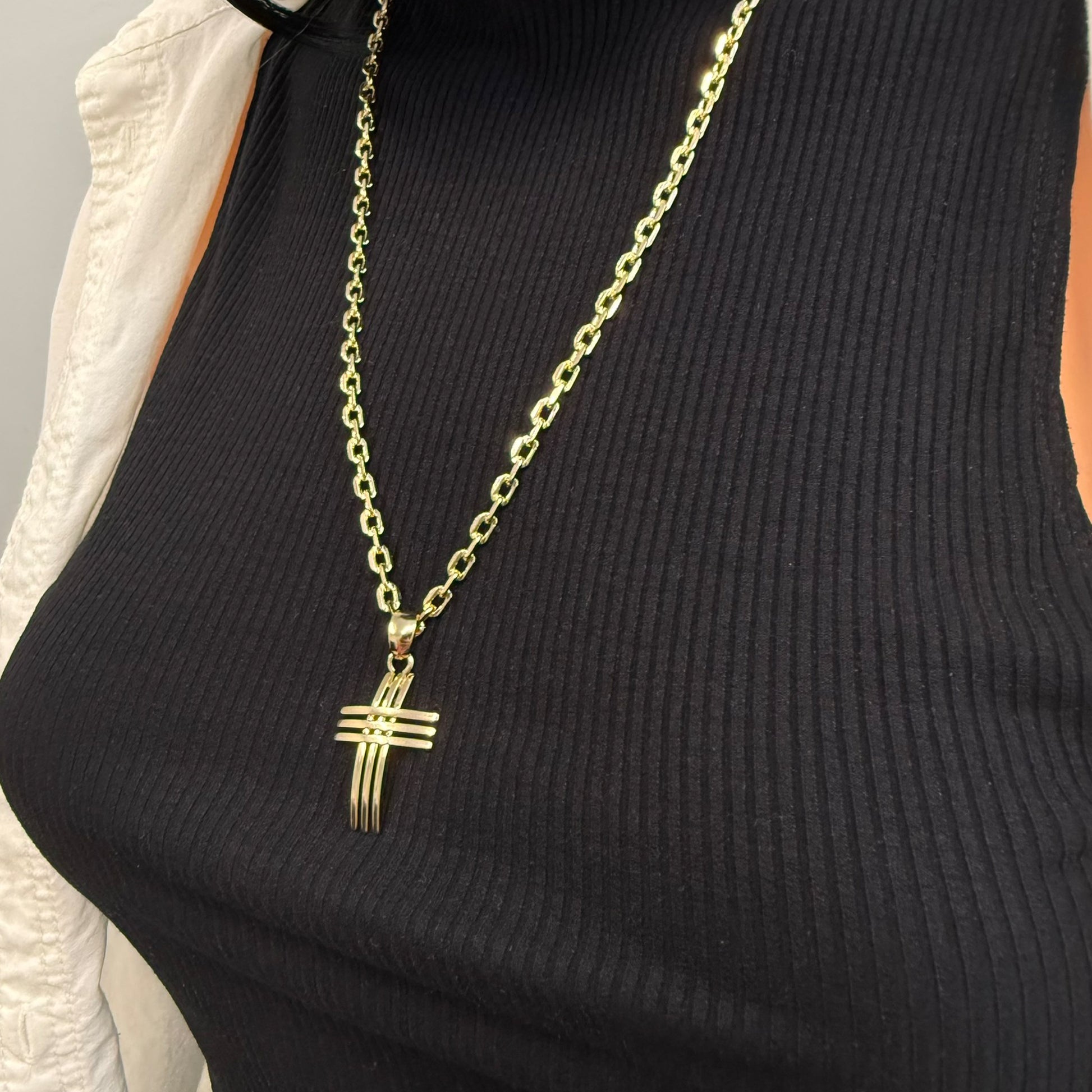 Close-up angled view of a mannequin wearing a bold 18k gold filled chain necklace with a curved dimensional cross pendant, styled over a ribbed black top and partially visible cream shirt.