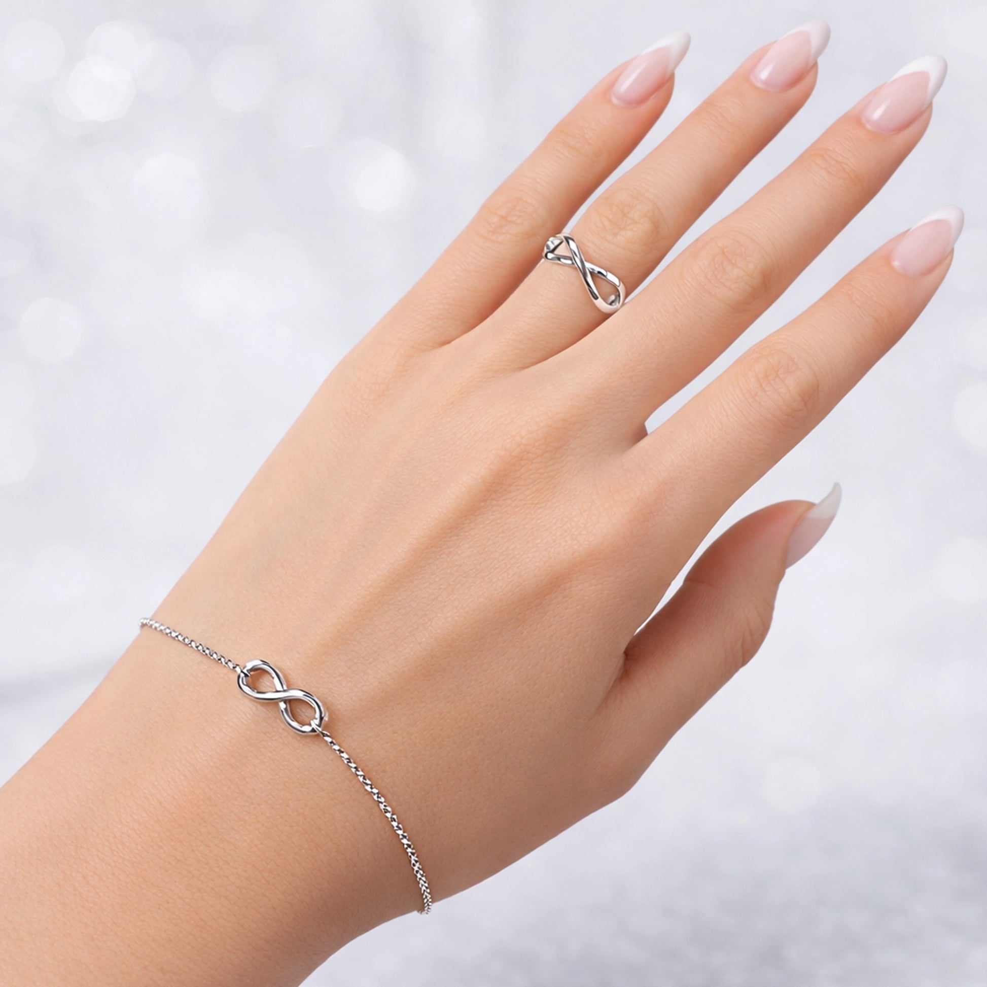 Sterling silver infinity ring and matching infinity bracelet on a slender hand, polished finish, minimalist jewelry set, French manicure, clean professional studio background.