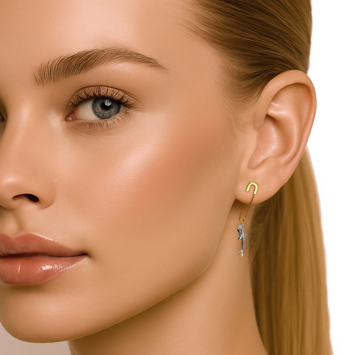 Half-profile close-up of a blonde woman with bright blue eyes, full makeup, glossy lips, and a centered ear wearing a unique dangle shark earring crafted in sterling silver with 18K gold plating. Clean white background and polished lighting highlight the edgy design.