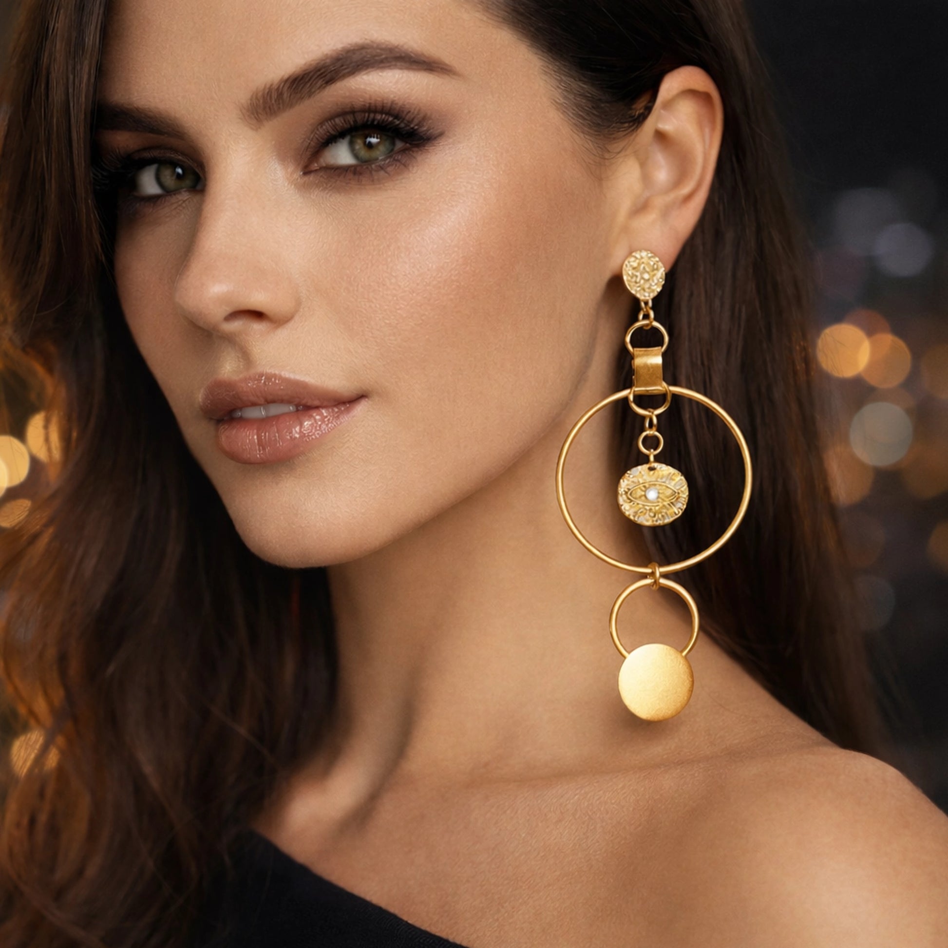 Elegant close-up portrait of a woman wearing statement gold drop earrings featuring textured stud tops, linked geometric rings, and dangling medallion charms, styled with soft glam makeup and warm ambient evening lighting in the background.