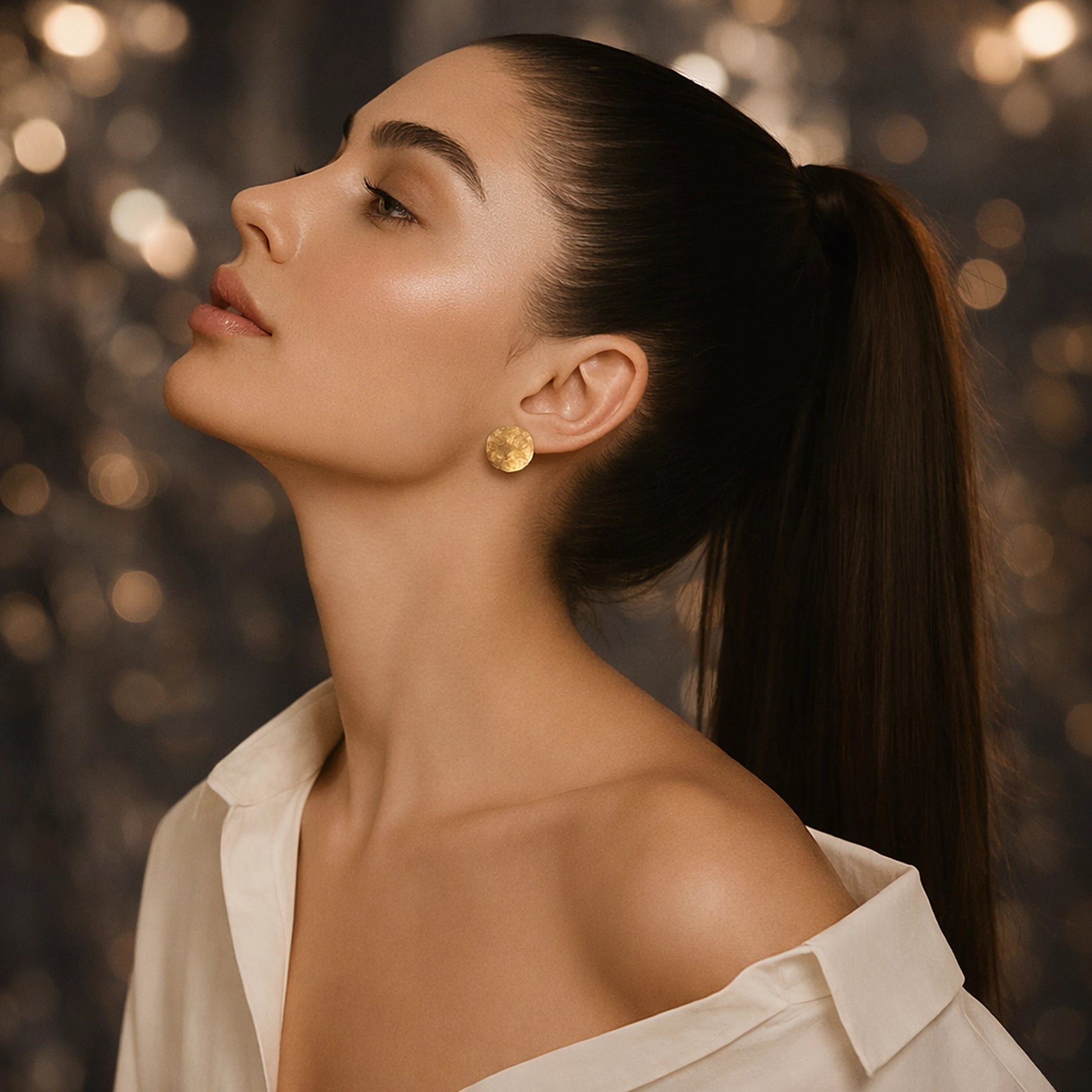 Elegant woman in an off-shoulder white blouse wearing simple round gold disc stud earrings, with her head tilted upward and hair in a sleek ponytail.