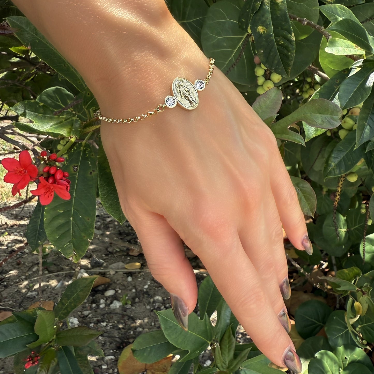 Religious gold bracelet showcasing a Lady of Grace medallion flanked by two sparkling CZs, styled on the wrist with vibrant garden greenery and red blossoms in the background. Dainty and meaningful gift idea.