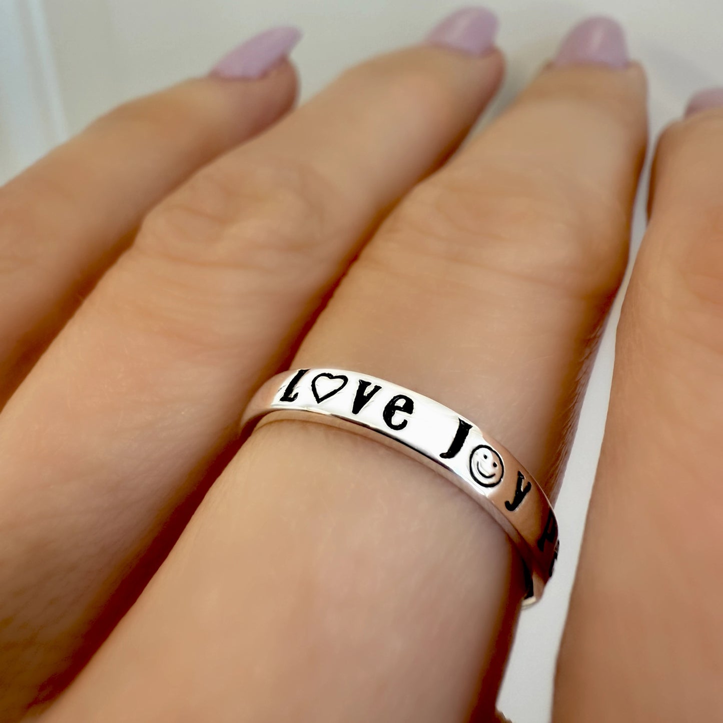 Sterling silver ring engraved with "Love", "Joy", and "Peace" using heart, smiley, and peace symbols, shown on a hand with pastel pink manicure.