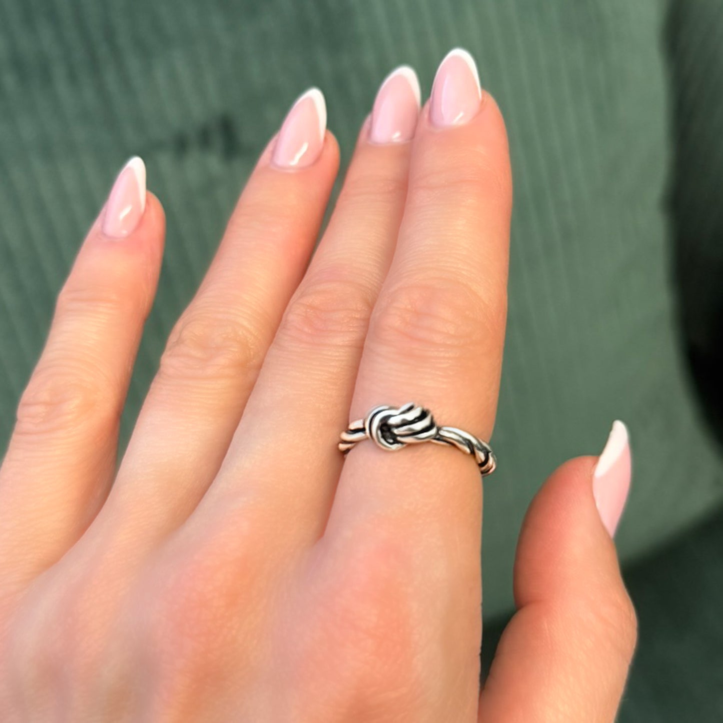 Close-up of a woman's hand wearing a sterling silver love knot ring with a twisted rope design, shown against a green fabric background. The nails are manicured in soft pink with white French tips.