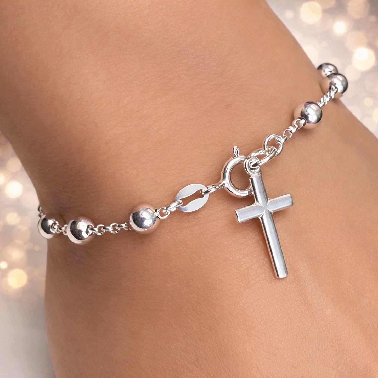 Close-up of a sterling silver ball beaded bracelet with a vertical polished cross charm dangling near the spring ring clasp. The bracelet is worn on a woman's wrist and photographed in soft, natural lighting.