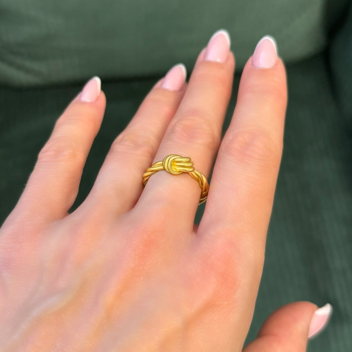 Elegant gold-plated sterling silver love knot ring on a woman's finger with light pink French manicure, photographed against a dark green background for contrast.