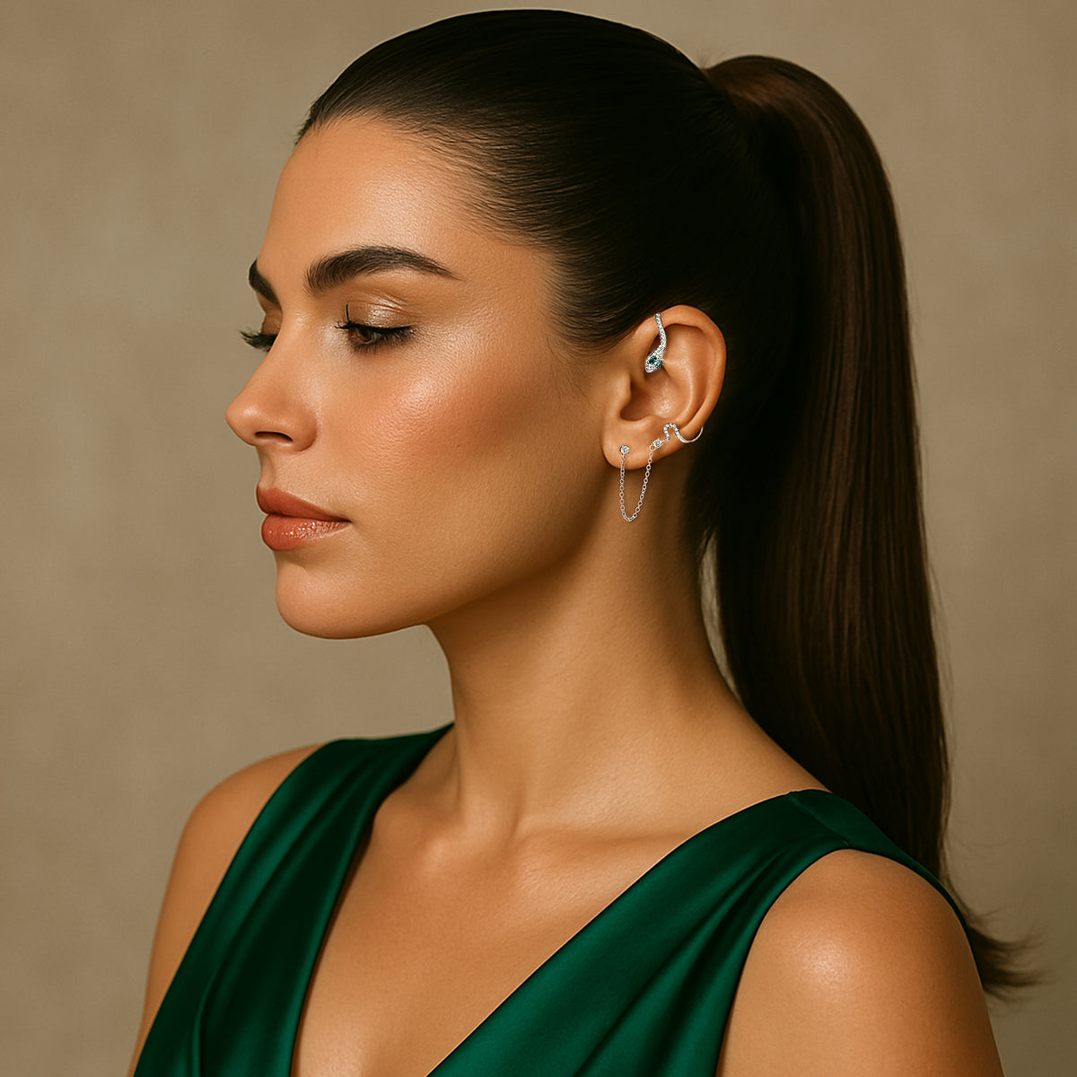 Woman with sleek ponytail wearing a stunning sterling silver snake cuff earring with green eyes and CZ stones. The cuff wraps around the upper ear and connects to a lobe piercing with a delicate chain. She’s dressed in an elegant emerald green dress against a neutral background.