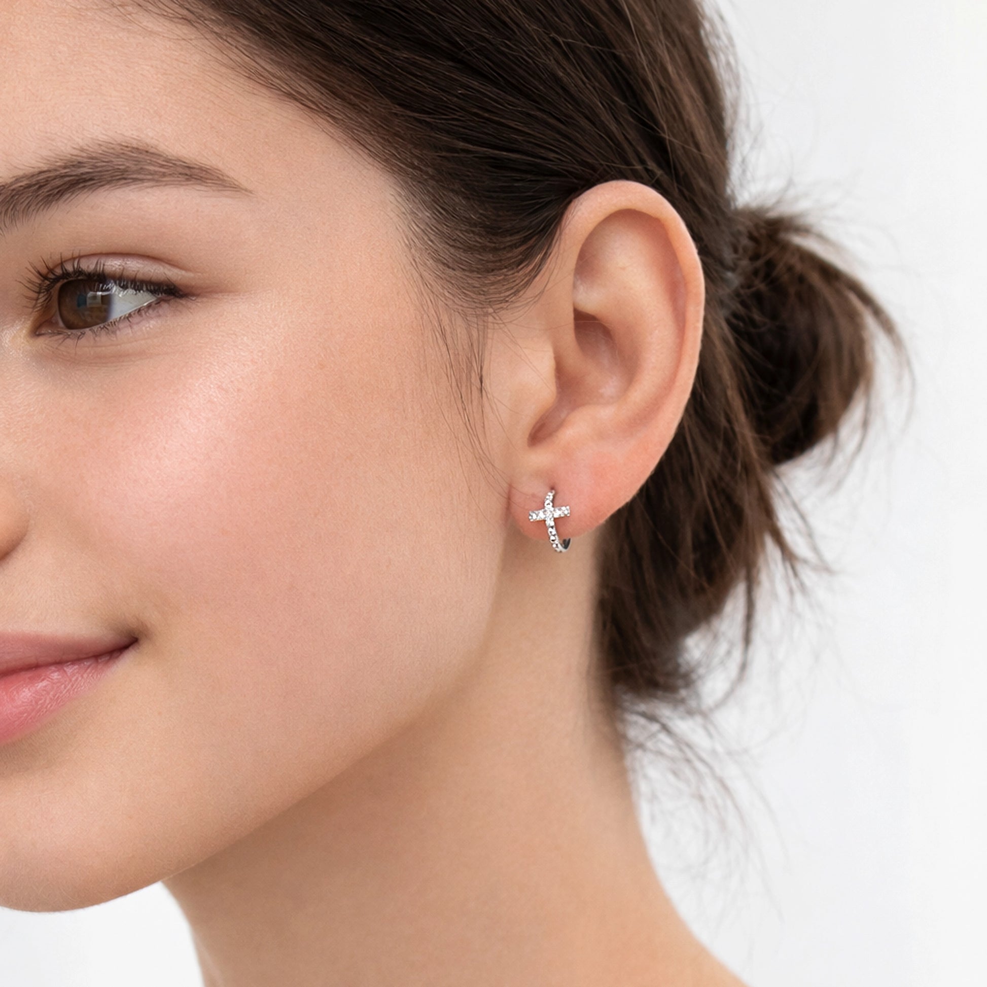 Tiny Cross Hoop Earring on teenage girl model, delicate silver cross huggie earring, minimalist Christian jewelry, small cross hoop earring close-up, elegant everyday religious earrings