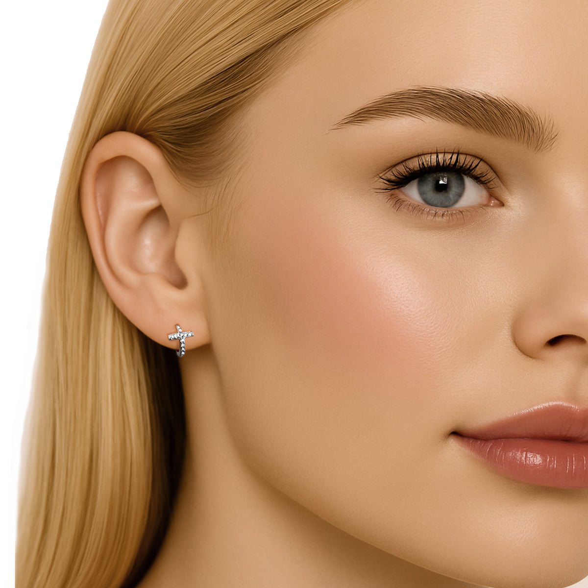 Half-profile close-up of a blonde woman with blue eyes, full makeup, glossy lips, and cheek highlighter. Her ear is centered and clearly visible, adorned with a tiny sterling silver CZ cross huggie hoop earring. Set against a clean white background.