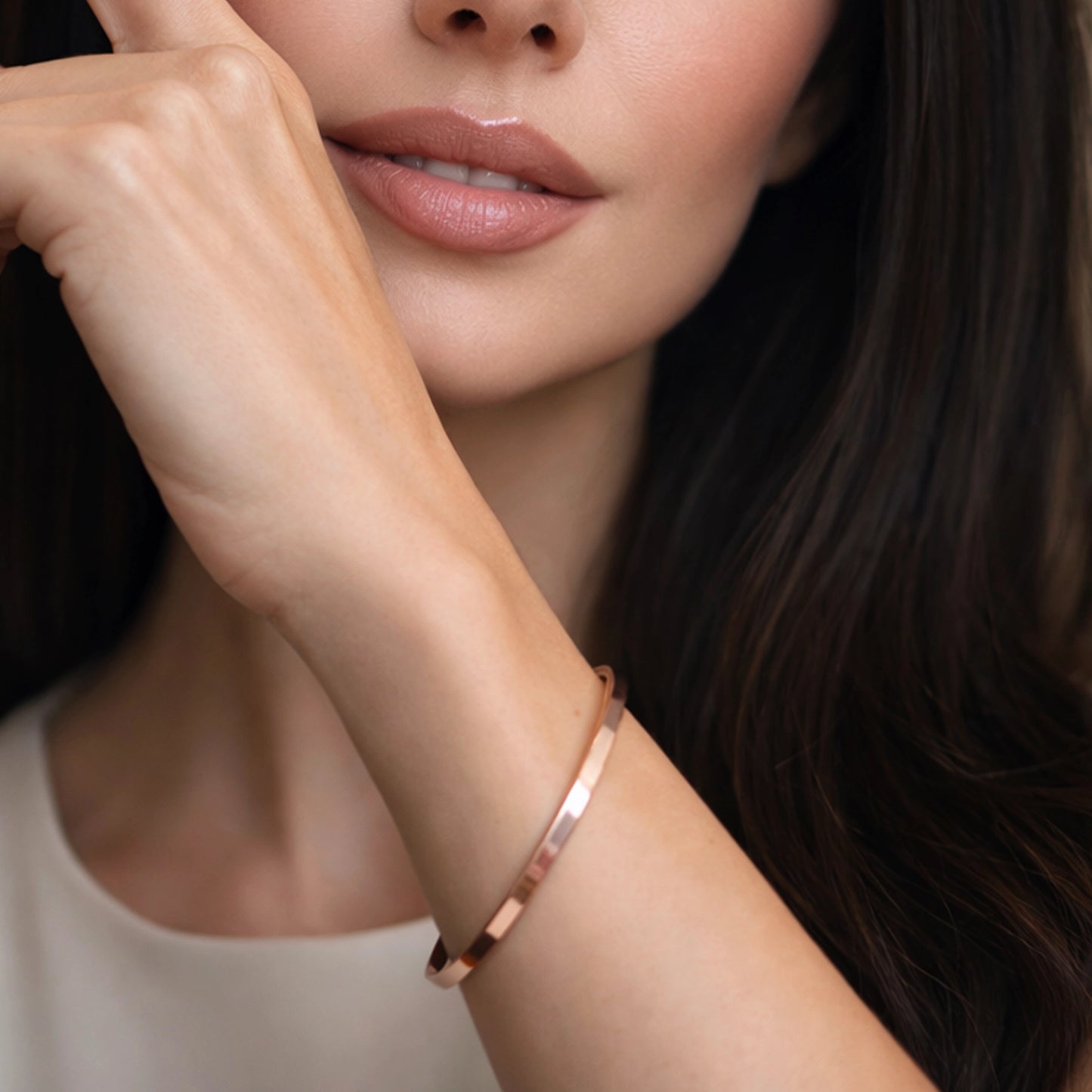 Beautiful elegant woman with flawless natural skin wearing a minimalist polished copper cuff bracelet on her wrist. Square close-up portrait with professional soft lighting and a warm blurred background. Focus on the copper cuff bracelet; no other jewelry visible.