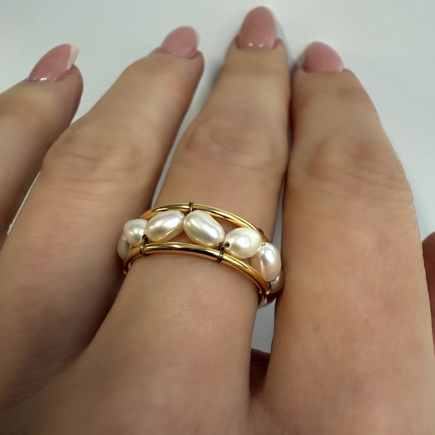 Hand wearing an adjustable freshwater pearl ring with double gold brass bands and white oval pearls, styled on manicured fingers with a soft white background.