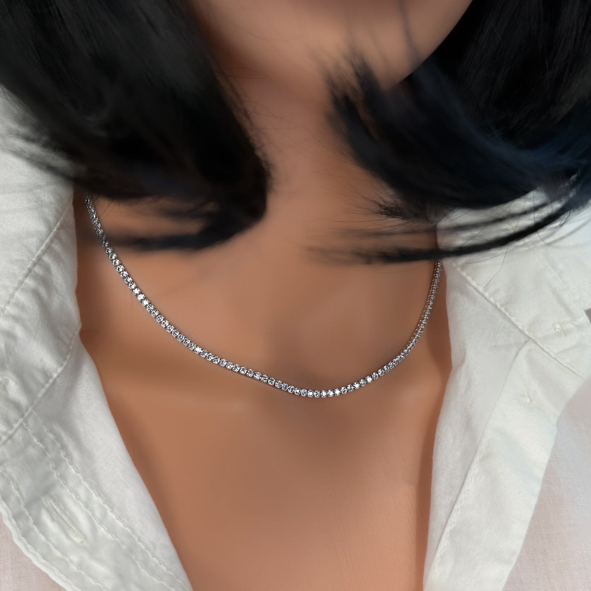 Close-up of a model wearing an adjustable sterling silver tennis necklace with a row of sparkling round cubic zirconia stones. Styled with an open white shirt and dark bob haircut, the necklace rests elegantly along the collarbone.